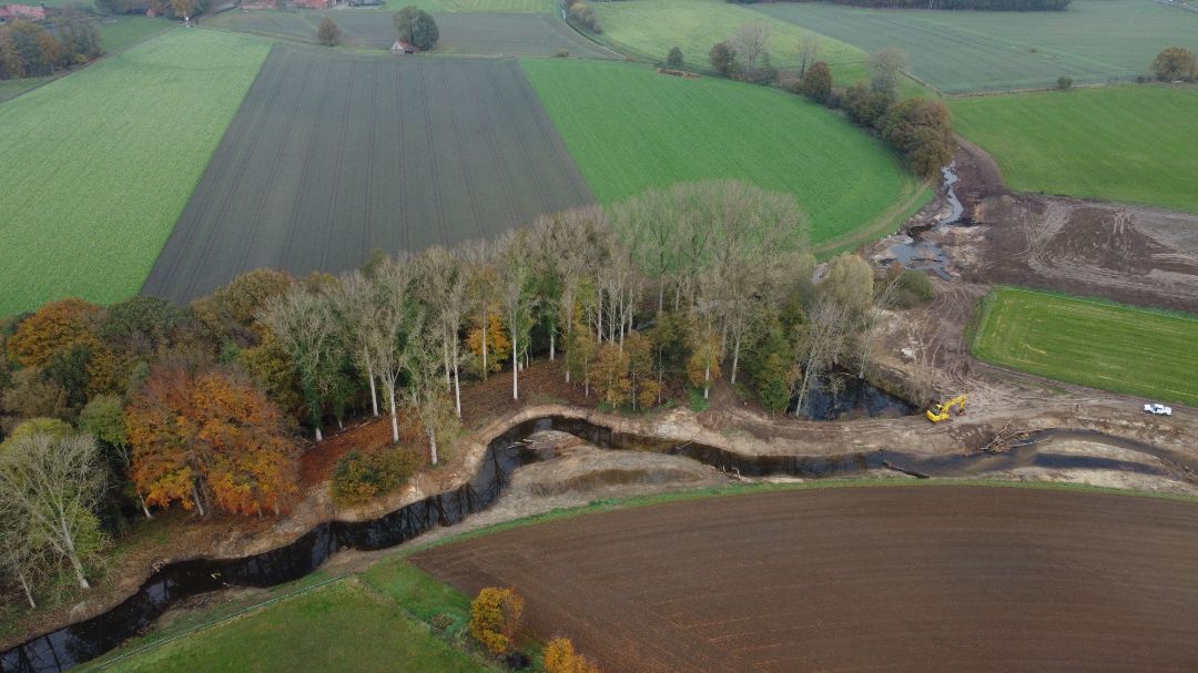 Blick im Herbst aus der Luft (Drohnenbild) auf ein renaturiertes, mit an einem Ufer mit B&auml;umen ums&auml;umtes Flie&szlig;gew&auml;sser sowie ein in dieses m&uuml;ndende kleineres Flie&szlig;gew&auml;sser.
Umgebend sind bewirtschaftete Felder zu sehen. 