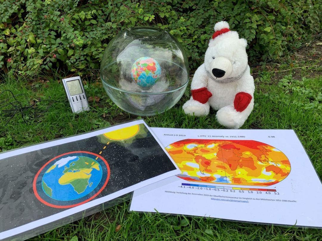 Materialien des Gruppenprogramms "Rette Dein Klima" im Maxipark: Ein Kuscheleisb&auml;r mit roter M&uuml;tze und roten Handschuhen sowie grauem Schal sitzt neben einer durchsichtigen Kugel, in der die Erdkugel zu sehen ist. Vor ihm liegen zwei eingeschwei&szlig;te Bl&auml;tter, auf denen Erde, Sonne und die klimatische Entwicklung auf der Erde abgebildet sind.