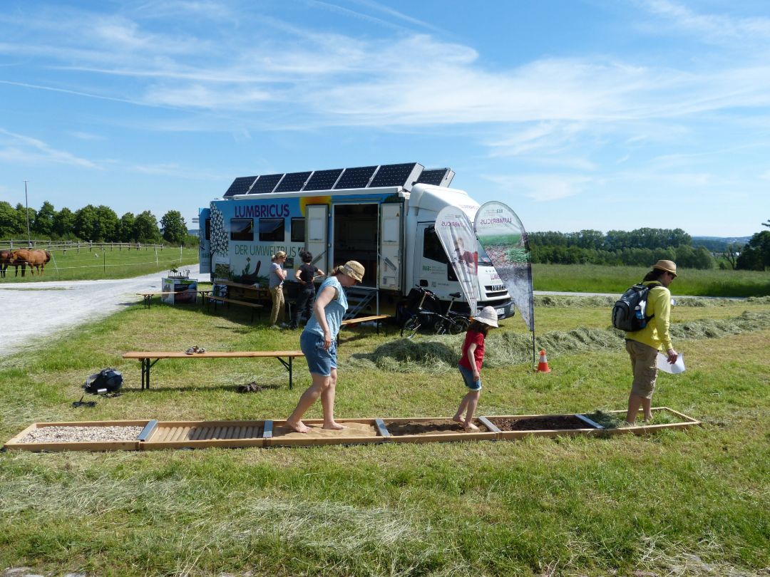 Blauer Himmel mit wenigen Wolken im Hintergrund.
Im Vordergrund ist ein Barfu&szlig;pfad aus Holz mit sechs F&auml;chern zu sehen, die mit unterschiedlichen Materialien (Holz, Steine, Sand...) gef&uuml;llt sind. Eine Frau, ein Mann und ein Kind gehen barfu&szlig; dar&uuml;ber. Dahinter steht der LUMBRICUS Umweltbus.