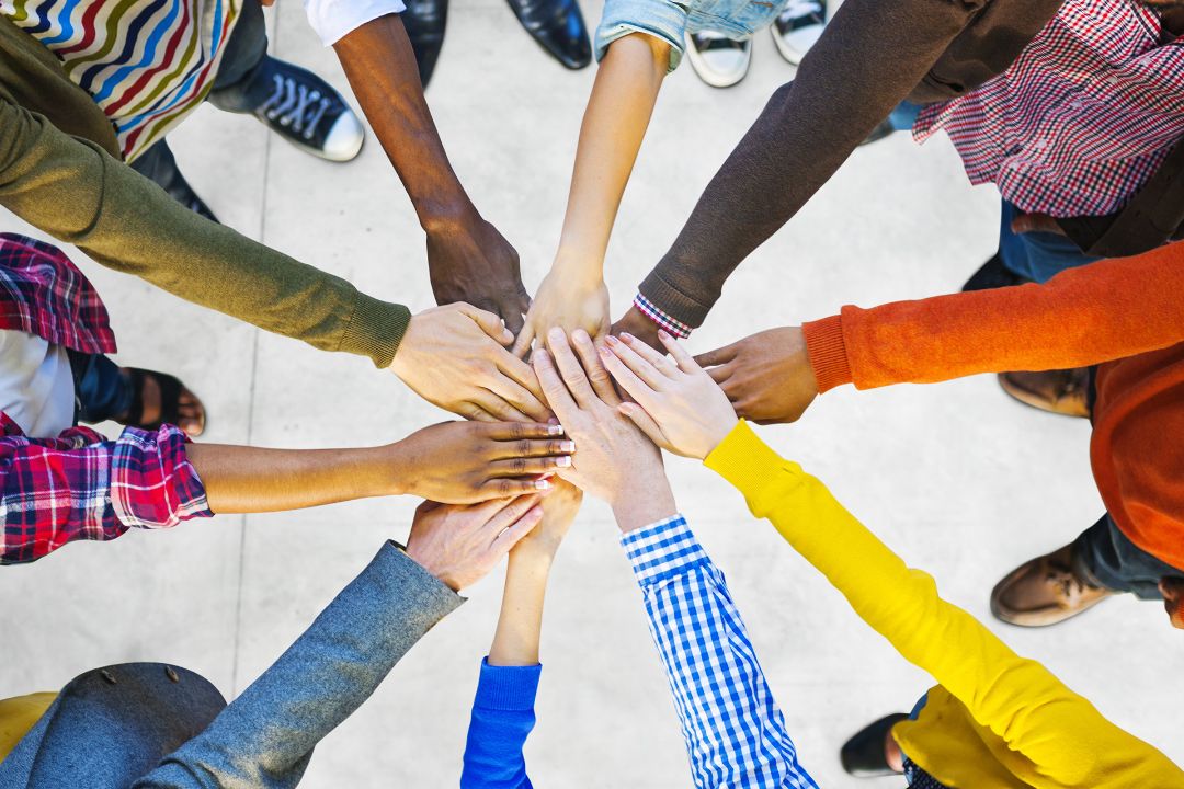 Menschen stehen im Kreis und strecken ihre H&auml;nde in die Mitte, wo sie sich sternf&ouml;rmig ber&uuml;hren.