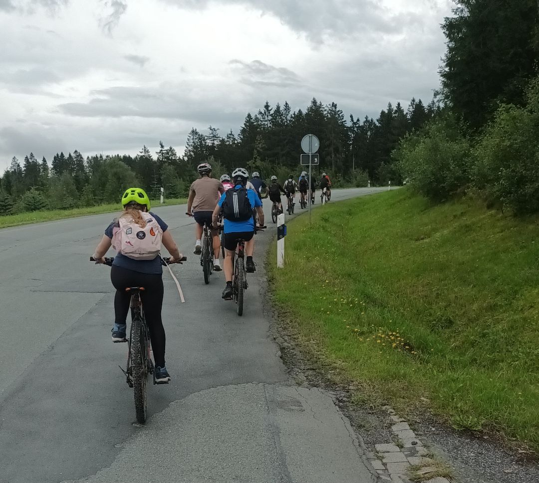 auf einer Landstra&szlig;e, die bergauf f&uuml;hrt, fahren in einer Kolonne hintereinander elf Radfahrer*innen mit Rucks&auml;cken auf den R&uuml;cken