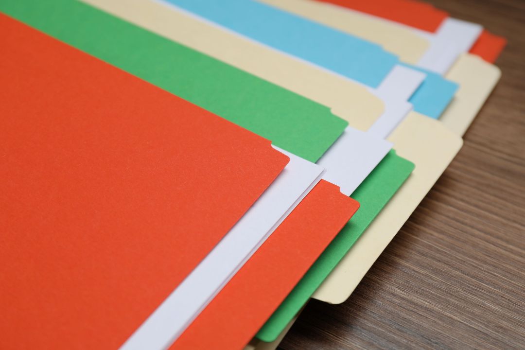 Sechs Papiermappen in den Farben orange, gr&uuml;n, creme und blau liegen aufgef&auml;chert auf einem braunen Holztisch.