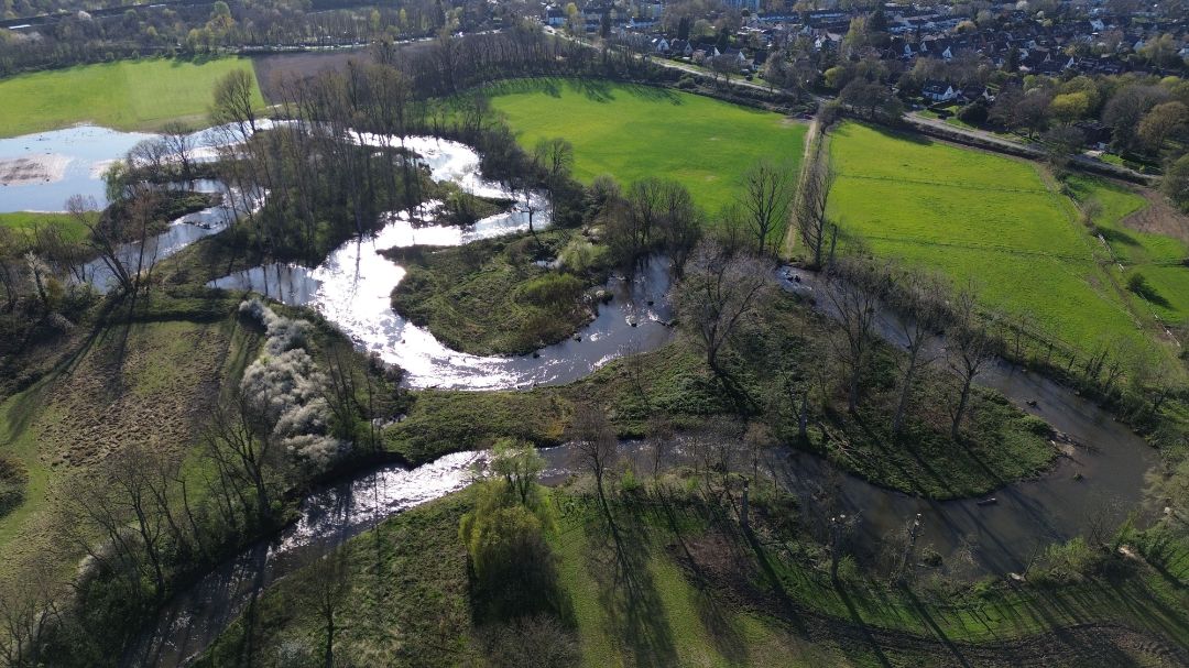 Blick von oben (Drohnenfoto) auf ein in mehreren Schleifen verlaufendes Flie&szlig;gew&auml;sser. An die Ufer angrenzend sind Wiesen zu sehen. Im Hintergrund befindet sich Wohnbebauung.