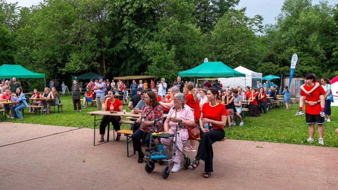 Festwiese vor der Veranstaltungsb&uuml;hne mit umliegenden Infost&auml;nden. Auf Bierb&auml;nken sitzen viele Menschen unterschiedlichen Alters und Geschlechts - gr&ouml;&szlig;tenteils in rote Shirts gekleidet.
