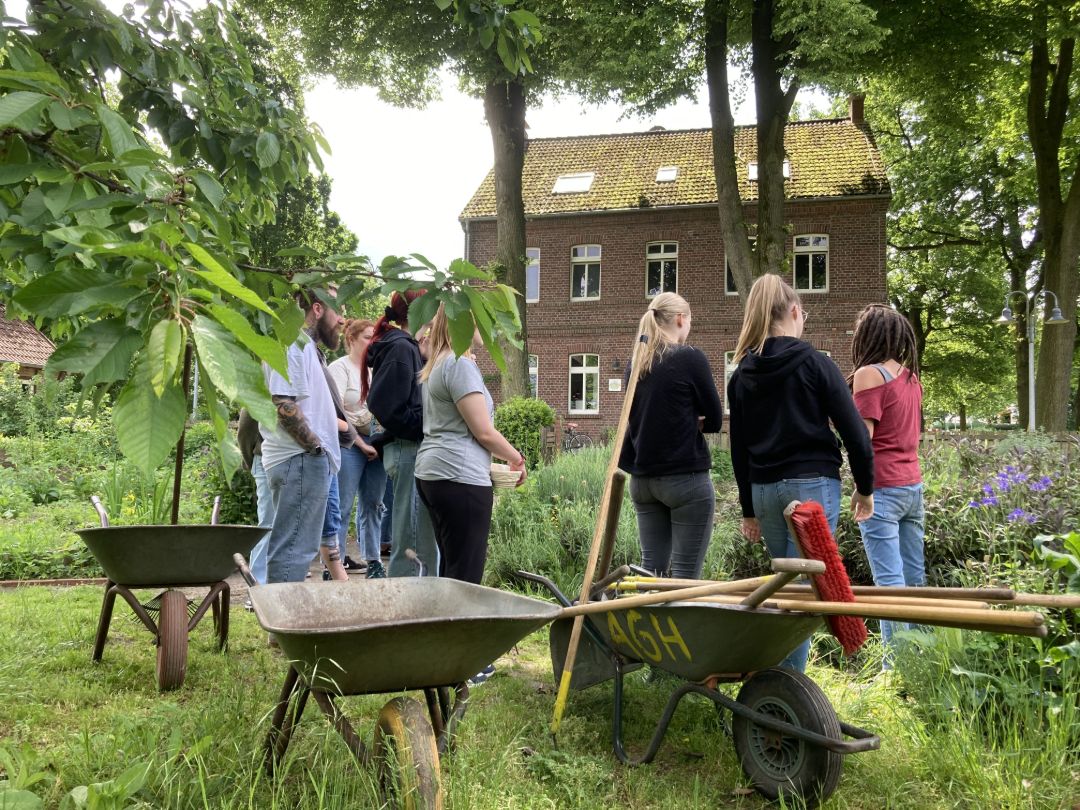 Mehrere Personen stehen hinter drei Schubkarren im Garten des Hof Wessels, der im Hintergrund zu sehen ist.