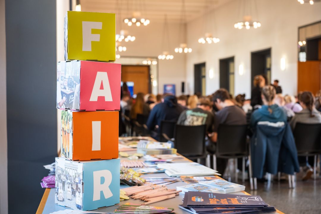 Jugendliche von Hinten in einem Veranstaltungssaal, im linken vorderen Bereich steht das Wort FAIR auf Pappw&uuml;rfeln, die auf einem Tische gestapelt sind