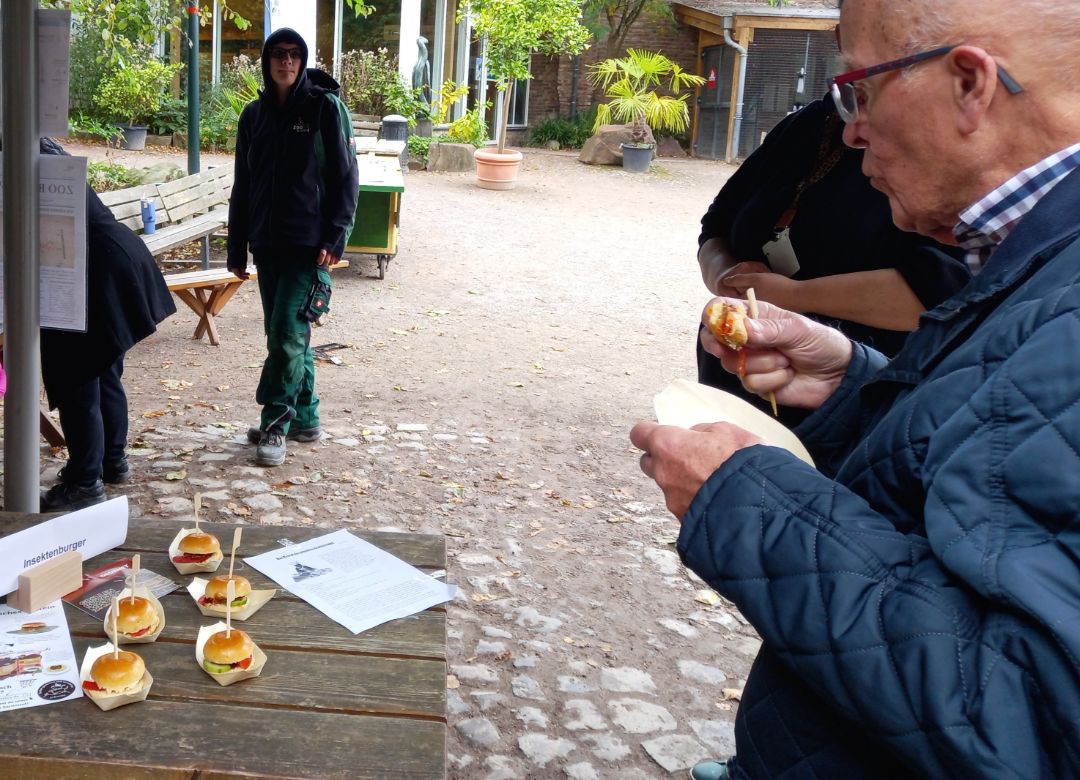 Ein Mann probiert einen Insektenburger. Dieser besteht aus einem kleinen Br&ouml;tchen und einer Frikadelle aus Insektenhackfleisch. Auf dem Tisch liegen weitere 5 Insektenburger.