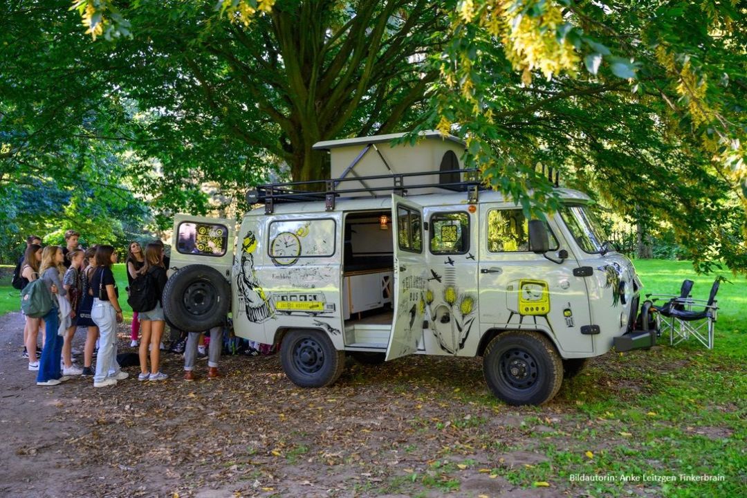 Eine Sch&uuml;ler*innengruppe schaut in den Zeitreisebus. Der Zeitreisebus parkt in einem Park unter einem gro&szlig;en gr&uuml;nen Baum. Im Hintergrund stehen 2 blaue Gartenst&uuml;hle. 