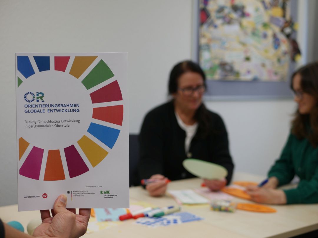 Die Publikation zum neuen Orientierungsrahmen f&uuml;r die gymnasiale Oberstufe wird hochgehalten. Sie zeigt das SDG-Wheel, ein buntes Zahnrad aus 17 Kacheln. Im Hintergrund arbeiten zwei Frauen mit Metaplankarten.