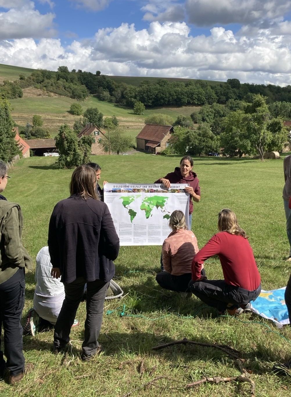 Eine Gruppe Lernender lauscht auf einem freien Feld in h&uuml;geliger Landschaft dem Vortrag der Referentin, die ein Plakat mit der Welt und der &Uuml;berschrift "Biologische Vielfalt und indigene V&ouml;lker bedroht" hochh&auml;lt.
