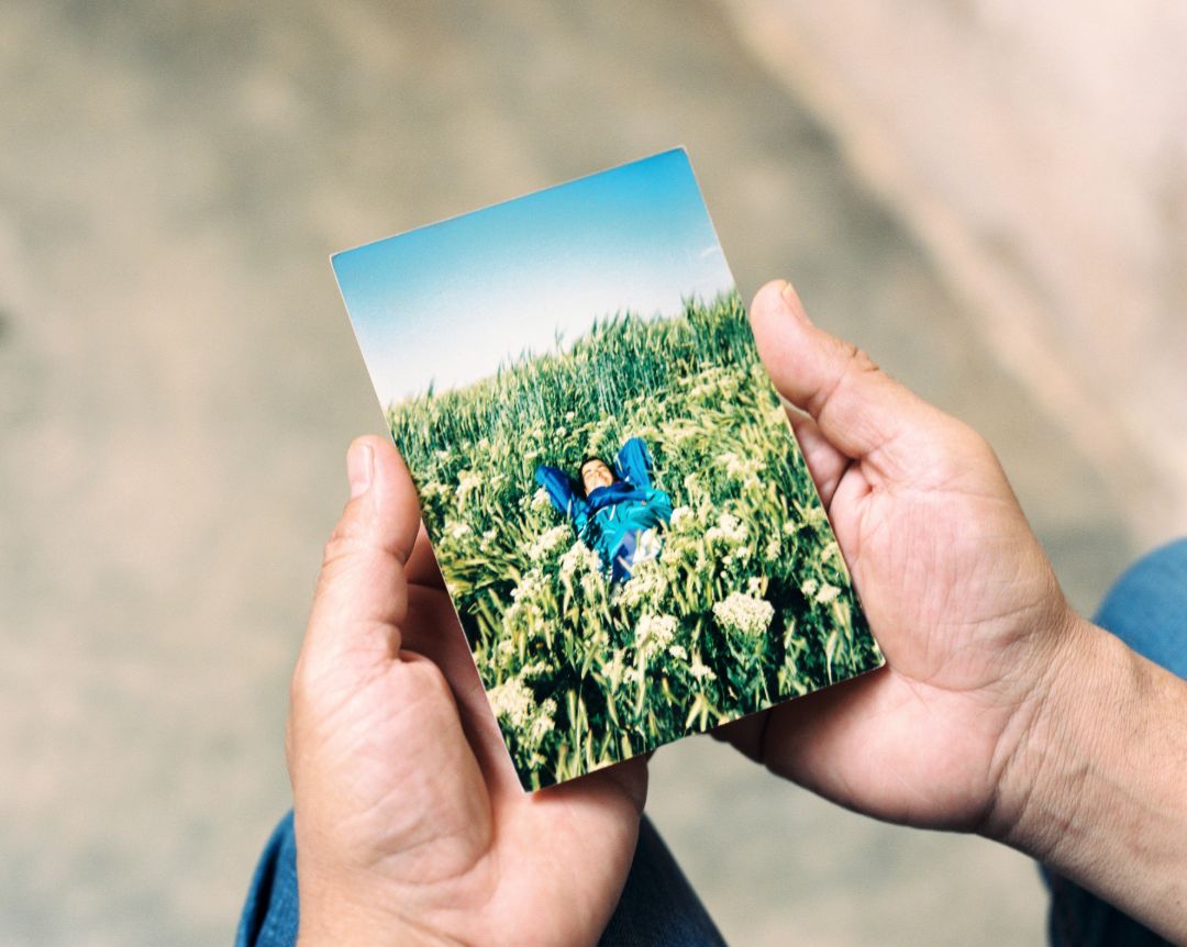 Das Bild zeigt ein Foto von einem gl&uuml;cklichen jungen Mann, der in einer bl&uuml;henden Wiese liegt. Das Foto wird von zwei H&auml;nden gehalten.