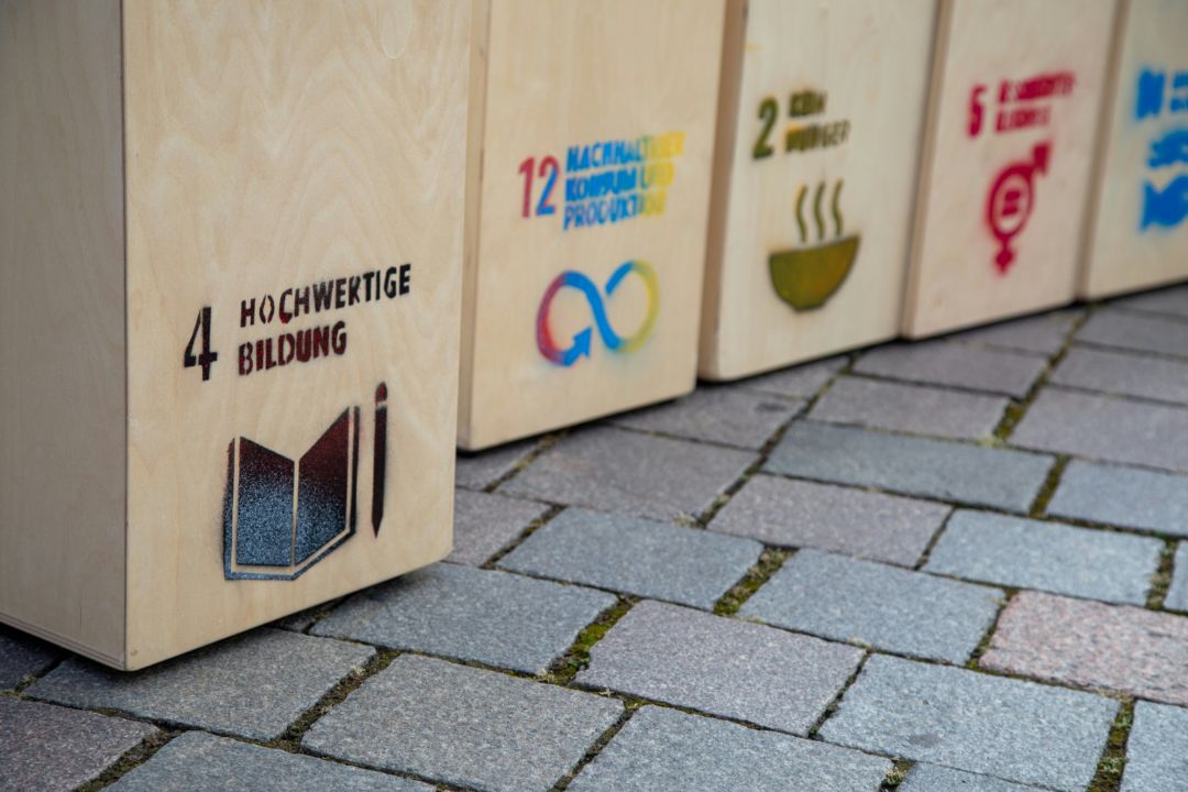 In einer Reihe befinden sich Holzk&auml;sten mit aufgedruckten SDGs auf einer gepflasterten Stra&szlig;e. Nummer 4, Hochwertige Bildung, ist vorne im Bild und vorgezogen.