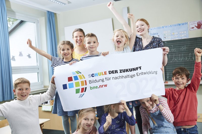 In einem Klassenzimmer hält eine Kindergruppe und zwei Lehrerinnen ein Plakat mit dem Logo hoch. Das Logo ist ein stilisierter Globus aus siebzehn bunten Kacheln und dem Schriftzug "Schule der Zukunft. Bildung für nachhaltige Entwicklung. bne.nrw".