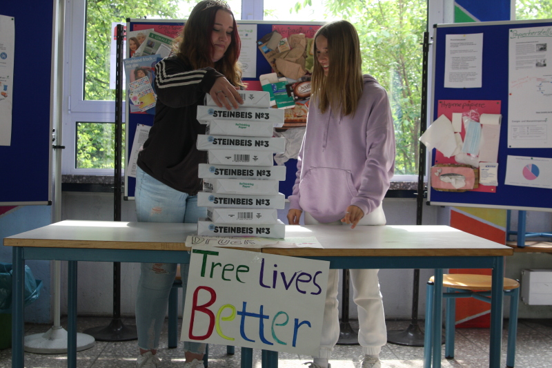 Zwei Mädchen an einem bunten Infostand zum "Tag ohne Kopien" stapeln Papierpakete übereinander.