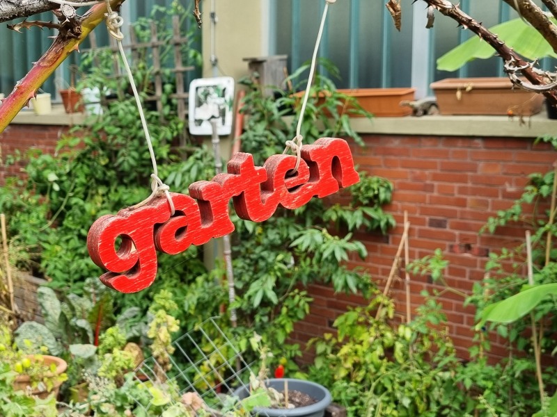 Roter Schriftzug Garten aus Holz aufgehängt vor einer grün berankten Backsteinwand