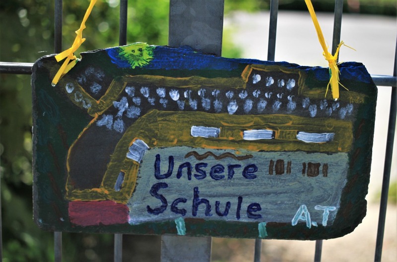 Eine Schieferplatte, auf der ein Kind mit bunten Farben seine Schule mit Außengelände aus der Vogelperspektive gemalt hat. Das Schild hängt an einem Metallzaun. Im Hintergrund sind unscharf grüne  Pflanzen und ein Weg zu erkennen.