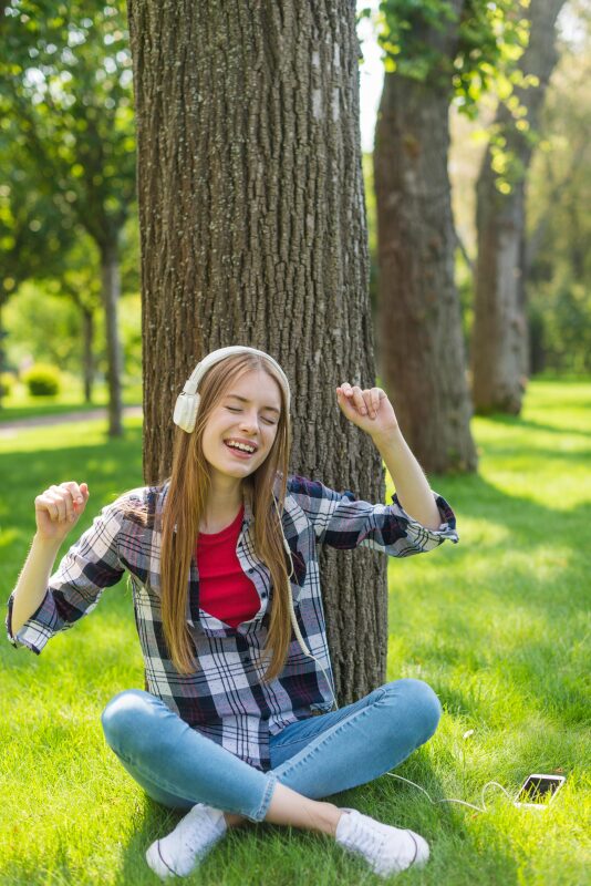 Eine Schülerin sitzt unter einem Baum und hört laut mit Kopfhörern Musik