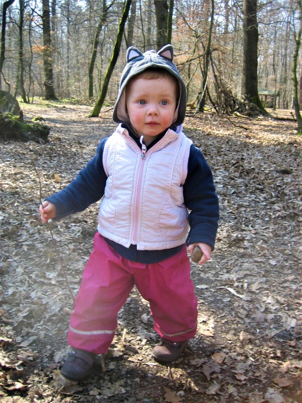 Ein etwa zweijähriges Kind steht in Matschmontur im Wald.